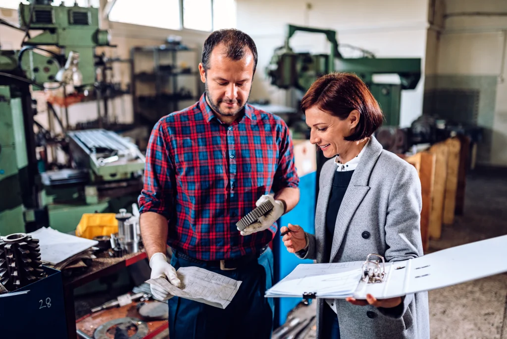 A man holding a metal part and paper stands next to a woman holding a clipboard in a workshop with industrial machinery in the background.